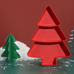A red silicone mold shaped like a Christmas tree, standing upright on a green surface with white faux snow and a small green paper tree prop against a solid dark red background.