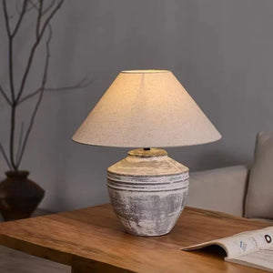 A rustic table lamp with a textured, distressed gray ceramic base and a warm beige fabric drum shade, shown lit on a wooden table in a cozy living room setting.