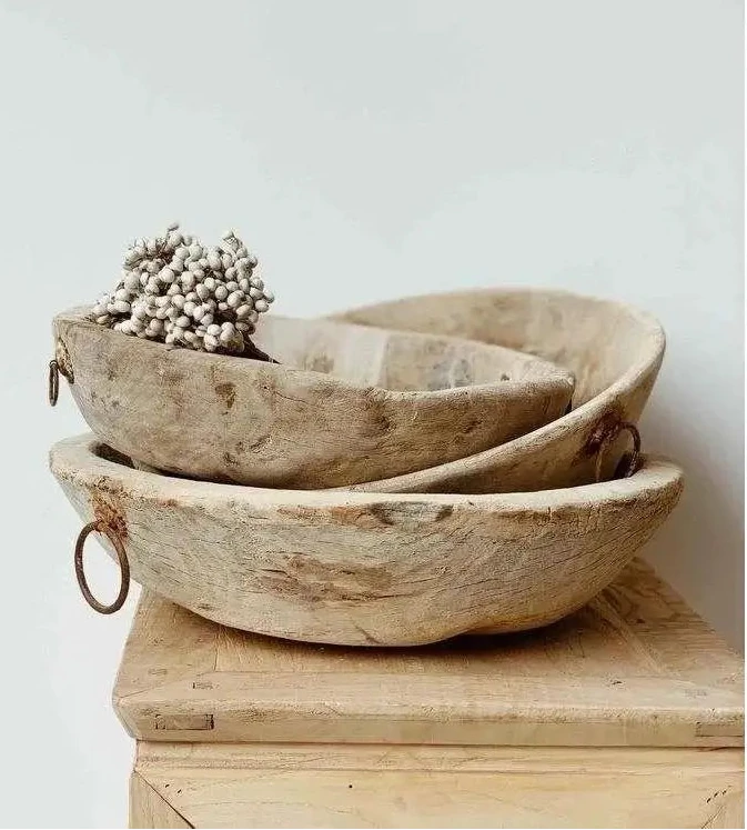 Three nested rustic wooden bowls with metal loop handles, displayed on a wooden block against a light gray background. The top bowl contains a small decorative cluster of dried flowers.