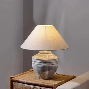 A table lamp with a warm beige fabric drum shade and a distressed gray ceramic base, shown lit on a woven rattan side table against a neutral gray wall.