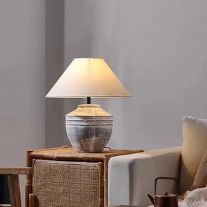 A table lamp with a textured, rustic gray ceramic base and a warm beige fabric drum shade, lit up and styled on a wooden side table next to a sofa.