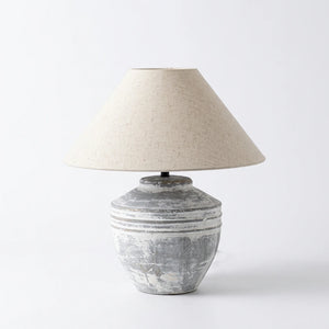 A rustic table lamp with a textured, distressed gray ceramic base and a warm beige fabric drum shade, shown lit on a wooden table in a cozy living room setting.
