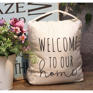 A cream-colored canvas welcome home doorstop with black text reading 'WELCOME TO OUR home' on a wooden surface next to a potted plant.