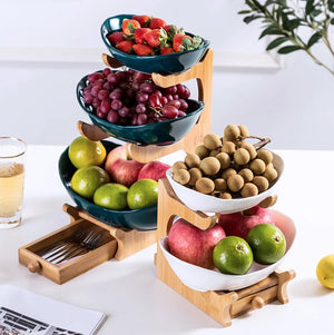 A three-tier bamboo and wood fruit stand holding bowls of strawberries, grapes, longans, apples, and limes on a white tabletop.