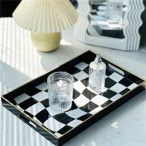 A rectangular black and white checkered tray with gold trim and side handles, styled on a light marble surface with a clear textured tumbler and a small bottle. A pleated cream table lamp and a mirror are softly blurred in the background.