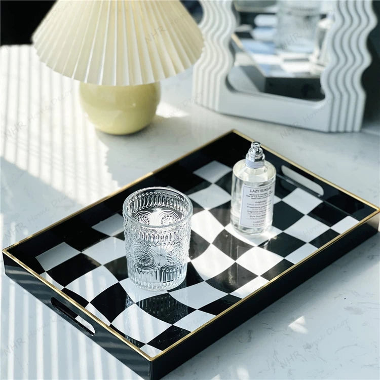 A rectangular black and white checkered tray with gold trim and side handles, styled on a light marble surface with a clear textured tumbler and a small bottle. A pleated cream table lamp and a mirror are softly blurred in the background.