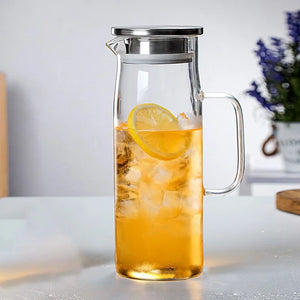 A clear glass pitcher with a stainless steel lid and handle, filled with iced tea and a lemon slice, set on a light gray countertop with a blurred kitchen background.