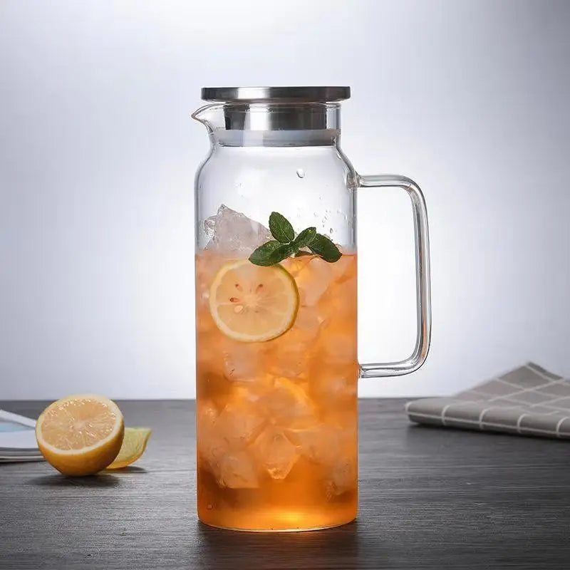 Clear glass pitcher with a metal lid and handle, filled with iced orange beverage, lemon slice, and mint leaves, set on a dark tabletop with lemon and a cloth in the background.