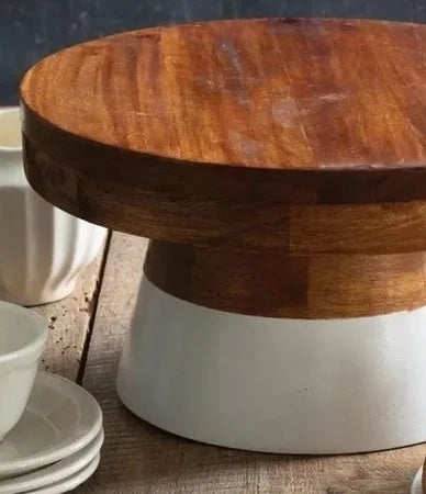 A round wooden serving board or cake stand with a thick white pedestal base, shown on a rustic wooden surface with white dishes partially visible in the foreground.
