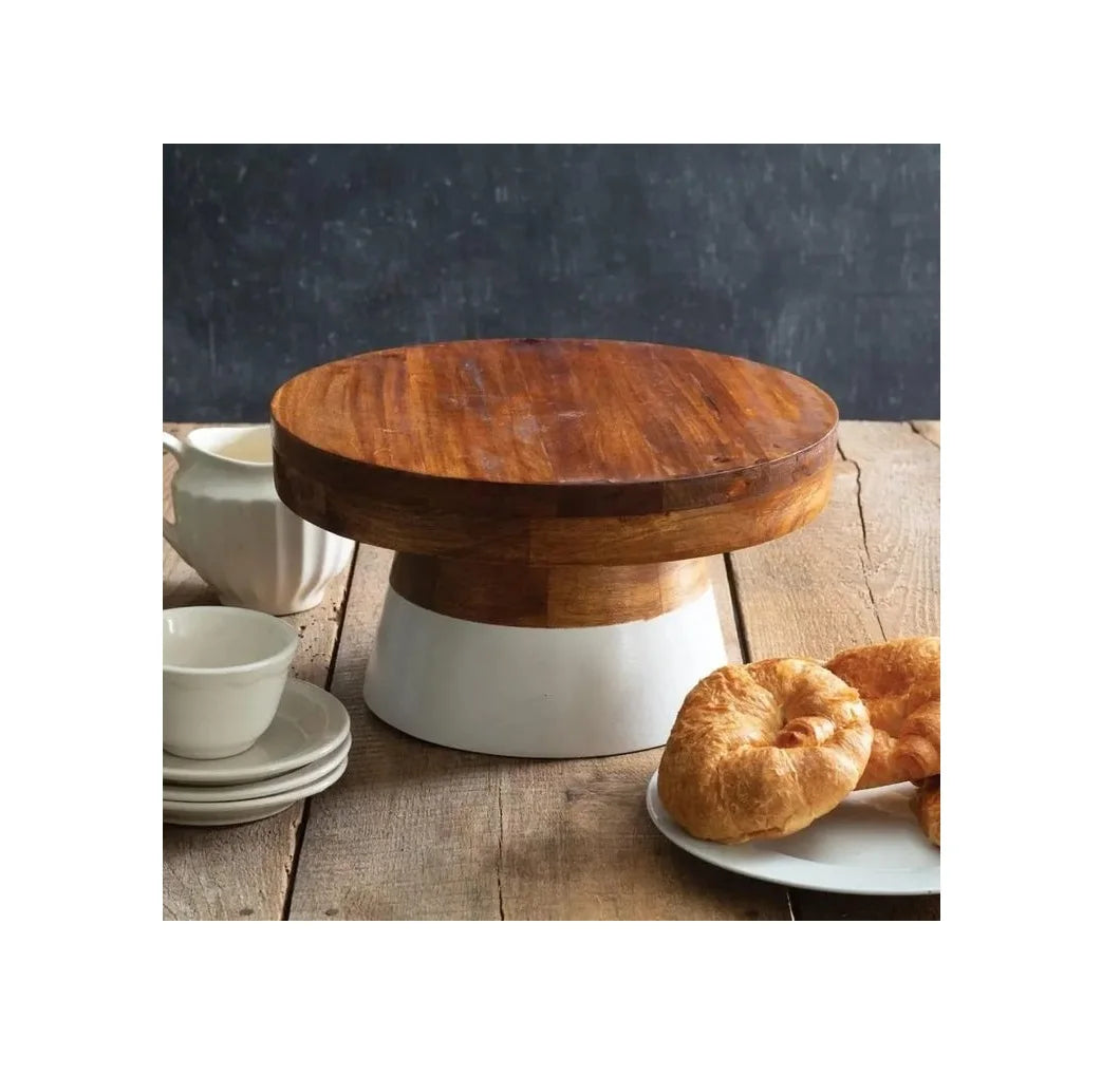 A round wooden cake stand with a white pedestal base, styled on a rustic wooden table with pastries and white ceramic dishes nearby.