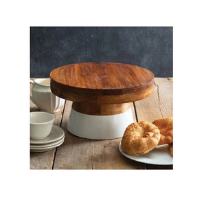 A round wooden cake stand with a white pedestal base, styled on a rustic wooden table with pastries and white ceramic dishes nearby.