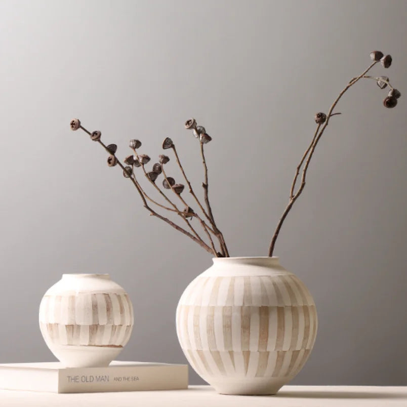 Two ribbed ceramic vases in warm off-white and beige tones, with the larger vase holding dried brown branches. One vase sits on a white book, and the other is placed beside it against a soft gray studio background.