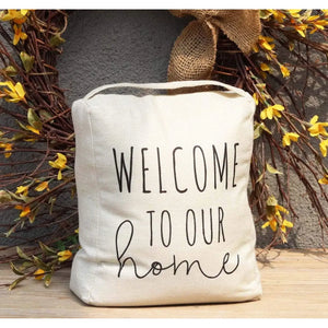 A square beige canvas sign with the phrase 'WELCOME TO OUR home' in black lettering, displayed in front of a decorative wreath with yellow flowers.